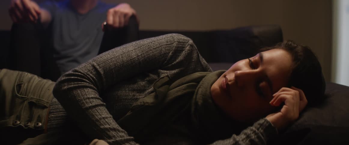 Woman with a head scarf lies down on a sofa in front of a man in the living room at home