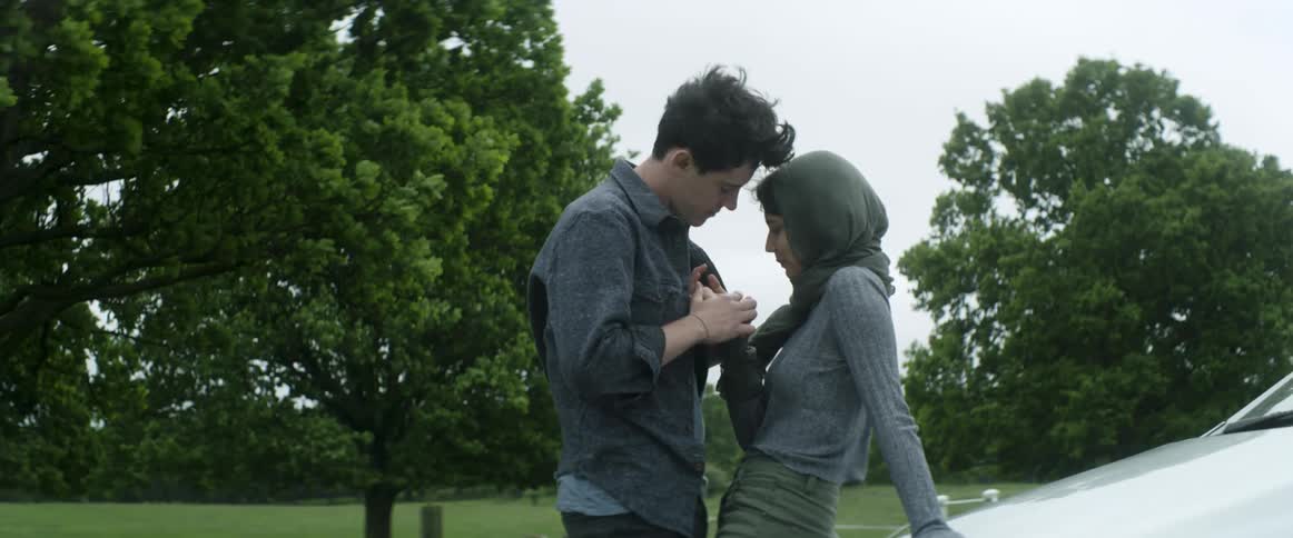 A teenager leans against a car in front of a park while her boyfriend holds her hands and leans close to her as he talks with her
