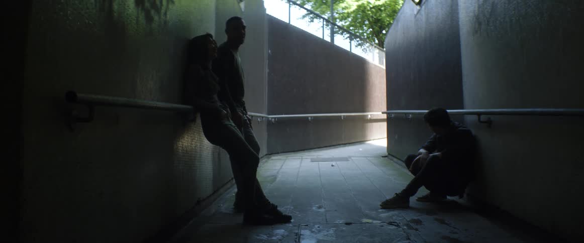 A woman and two men sit and stand in an outdoor tunnel with their heads hanged down