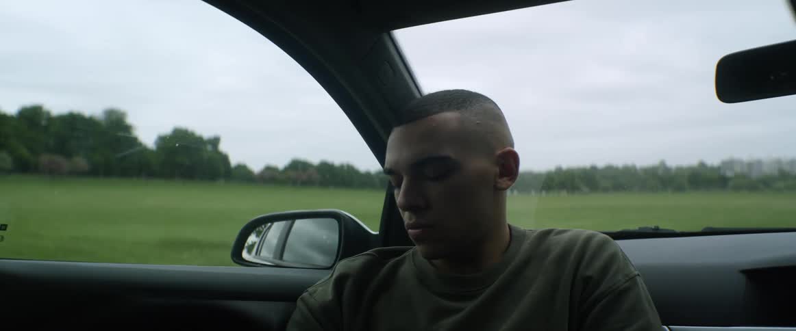 A man sits backward in a parked car in a field near a forest