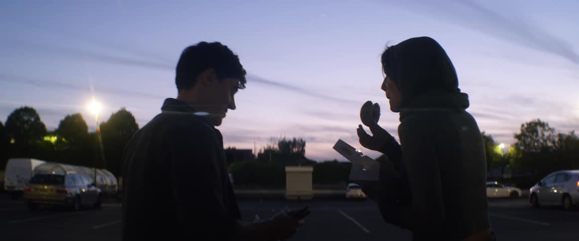 A woman and man chat while eating donuts in a parking lot at sunset