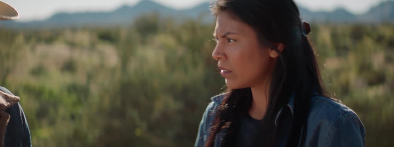 A man with a cowboy hat talking to a woman with landscape mountains and the sky in the background