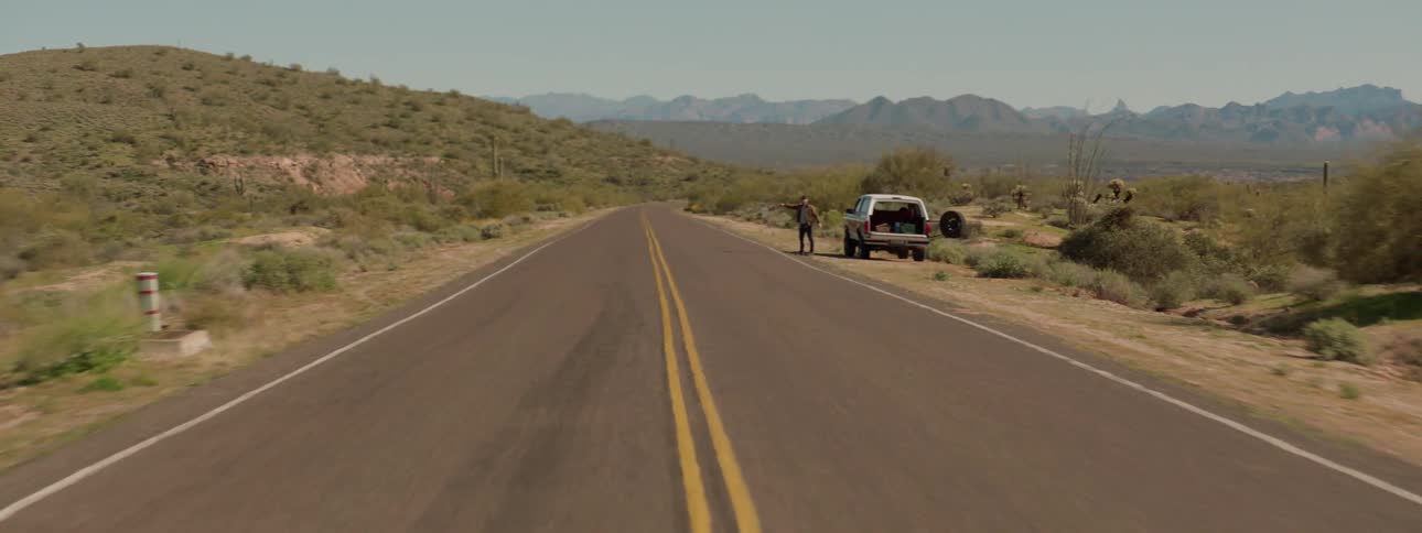 A man standing next to a broken down vehicle on a rural highway waves down a passing car for help