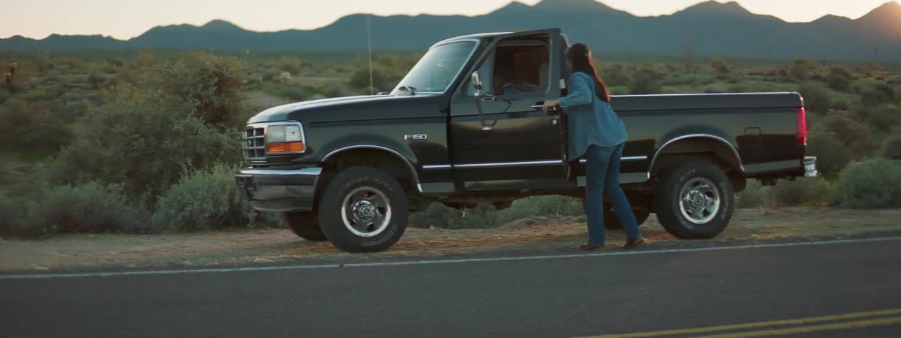 Two men change a tire on a vehicle as a teenager talks with a young girl in a truck