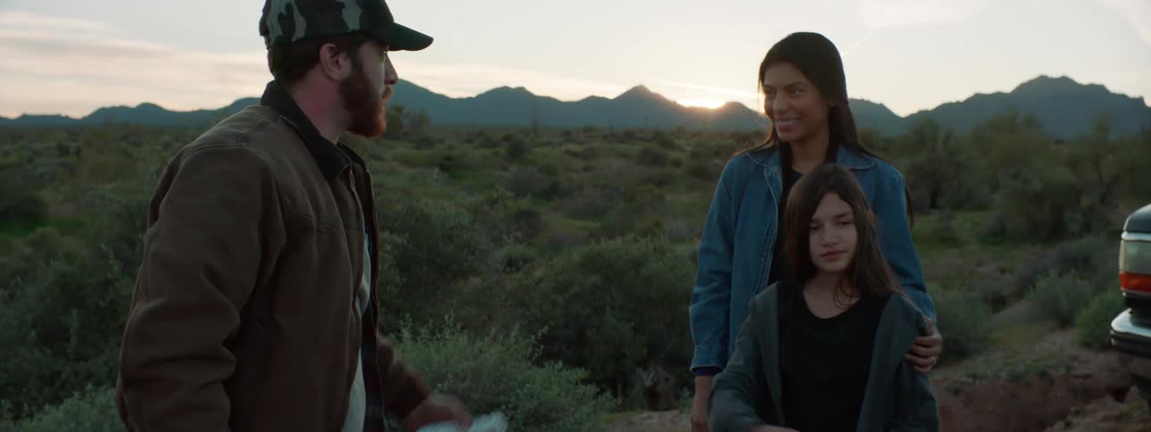 A girl hands food wrapped in aluminum foil to a man in a baseball cap before another man approaches to shake the mans hand