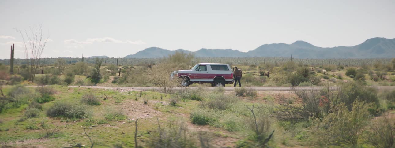 Person gets out of car to fix flat on highway cutting through scrub near mountain