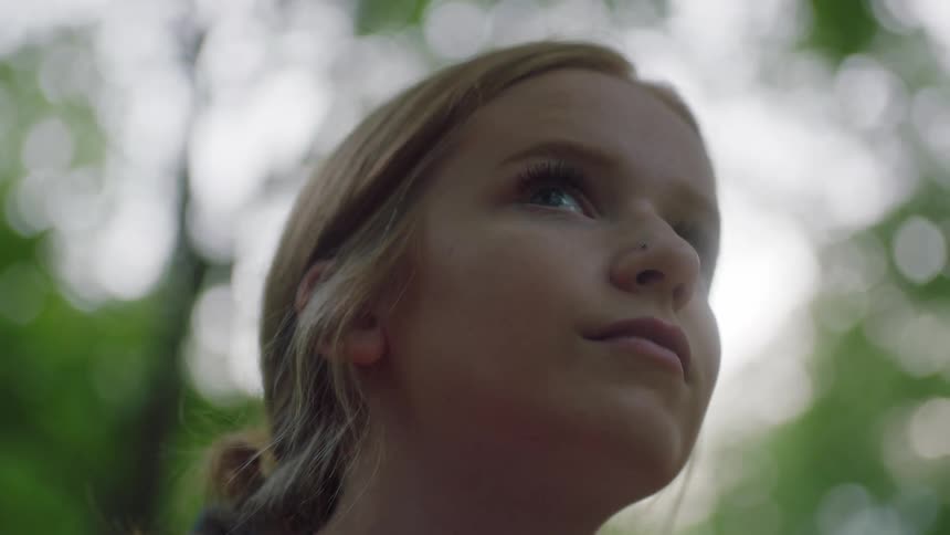 Girl with red hair sitting in a red hammock looks up at tall trees and turns her gaze to the camera in the forest