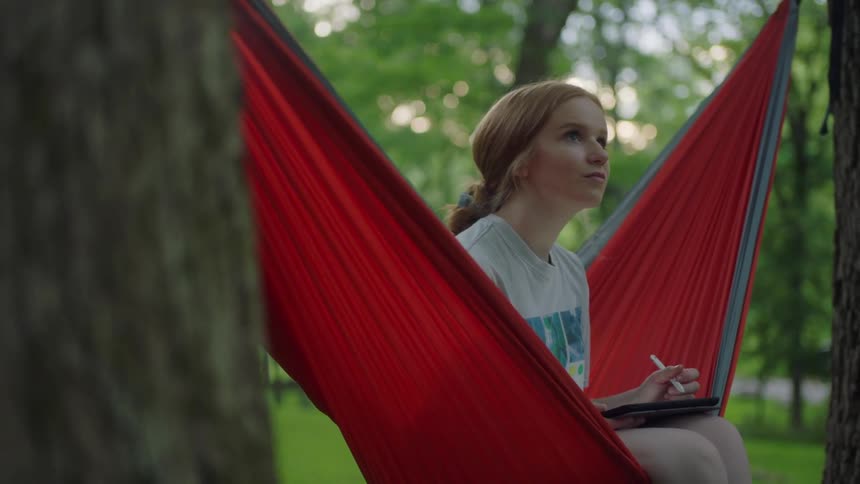 A young girl sits in a hammock outside and writes on a tablet