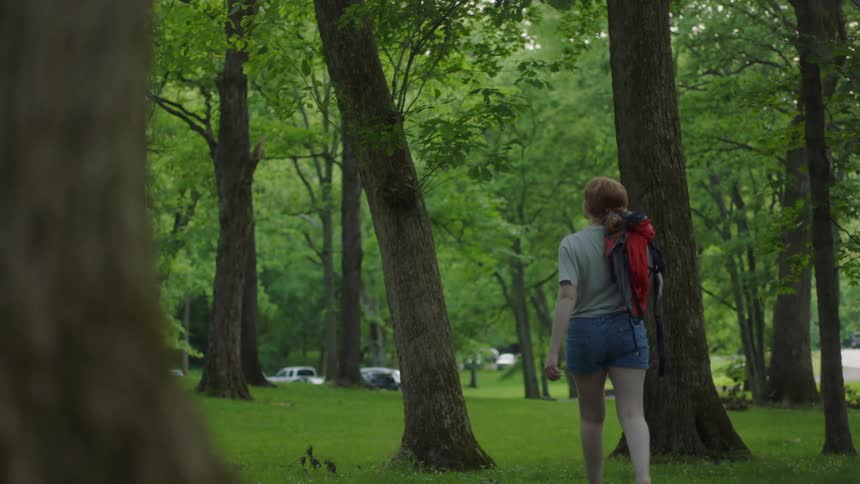 A woman walks through a treed park towards cars in the parking lot