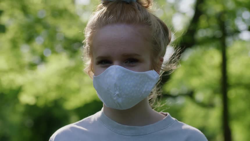 Girl with red hair and a light blue face mask looks at the camera in a forest