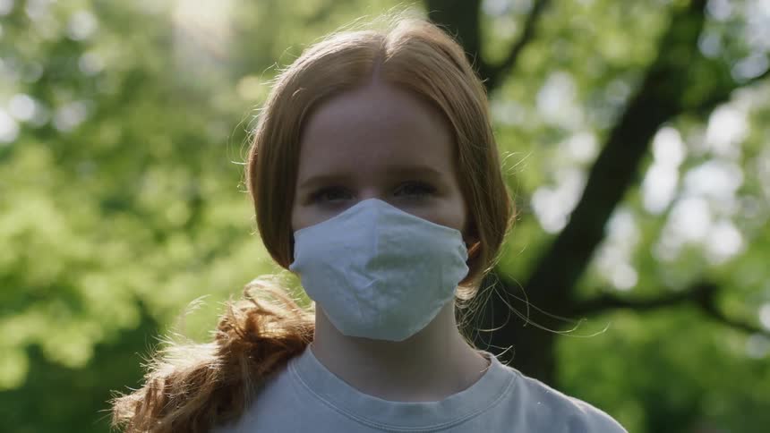 Girl with red hair and face mask raises her head to look at the camera in the forest