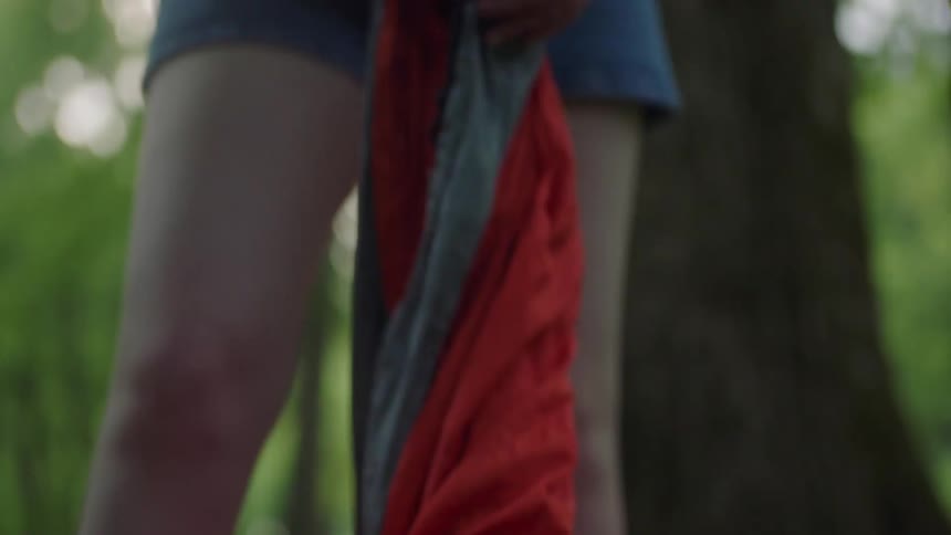 Girl with shorts and a grey t shirt sets up a red hammock in the forest