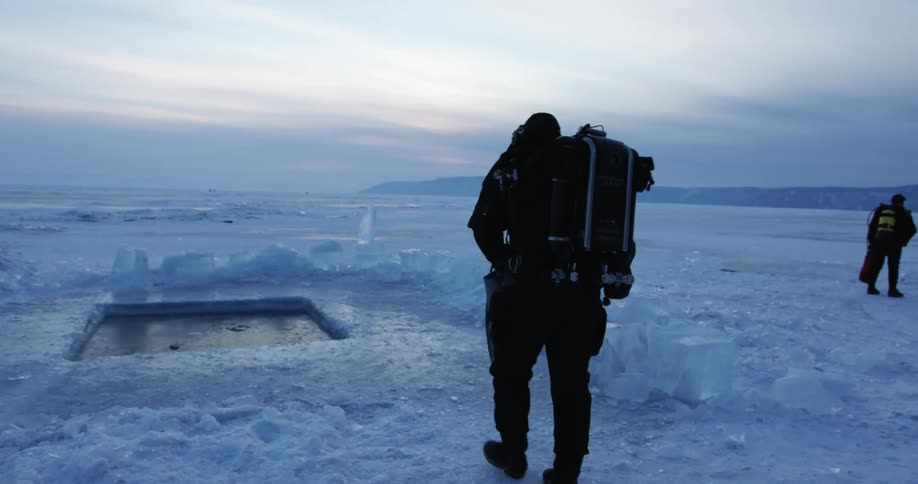 A researcher in the arctic carries flippers as he walks toward a square hole cut into the ice