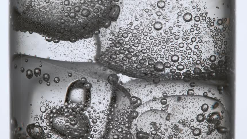 A glass is filled with a carbonated beverage and the small bubbles navigate around a number of ice cubes in the liquid
