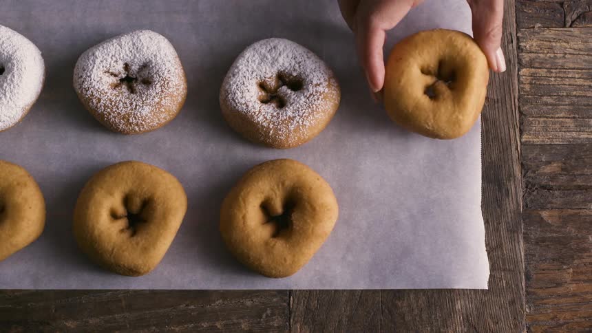 A hand places a doughnut on baking paper over a wooden table