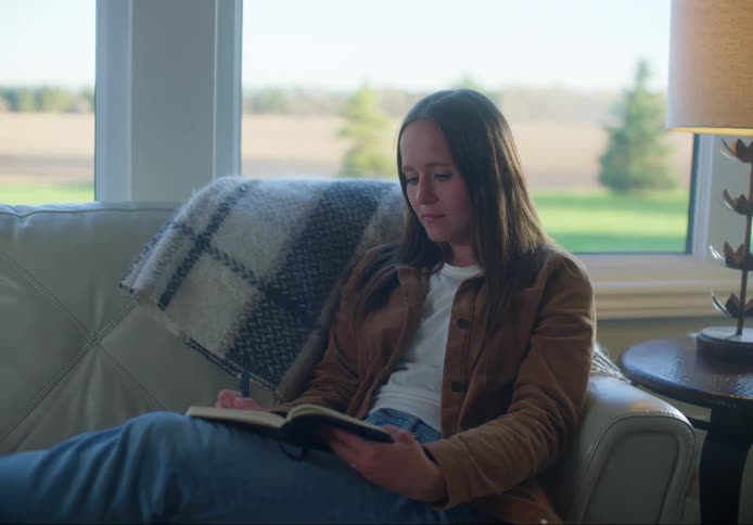 Woman with brown straight hair and jeans sitting on a sofa writes in a notebook in the living room