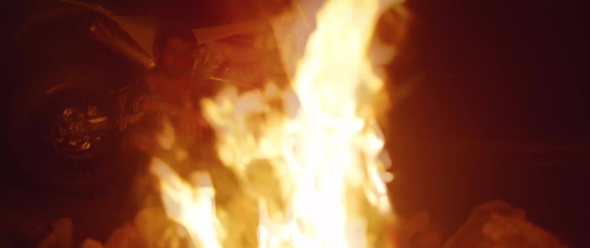 A man leans against a motorcycle as he sits in front of bonfire