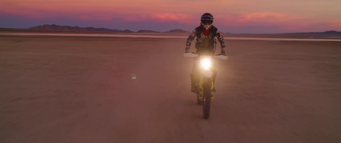 Motorcyclist rides through desert backed by red sky and mountains