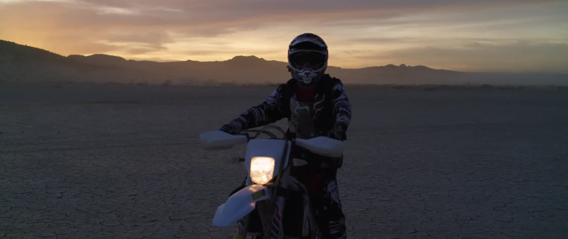 A person wearing crash helmet sitting on a stationary motorcycle in the desert at sunset