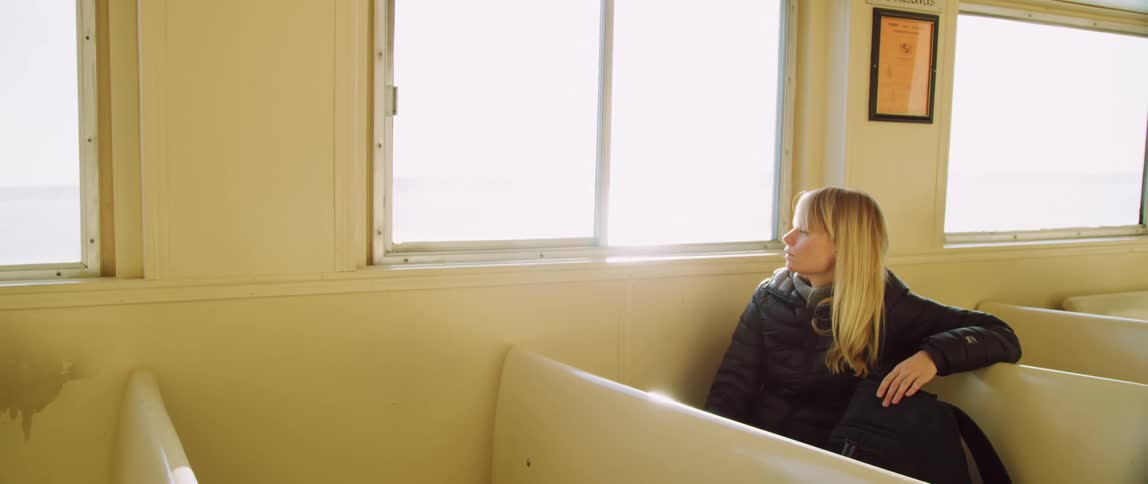 A woman taps her backpack while looking out a window of the passenger ferry she is traveling on