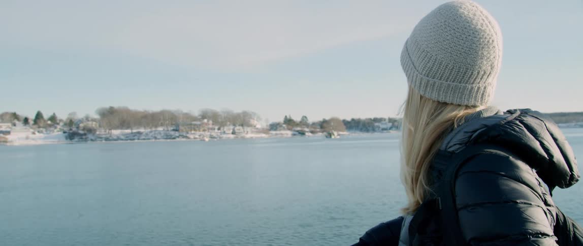 A woman stands at the bow of a ship observing the approaching coastline covered in snow