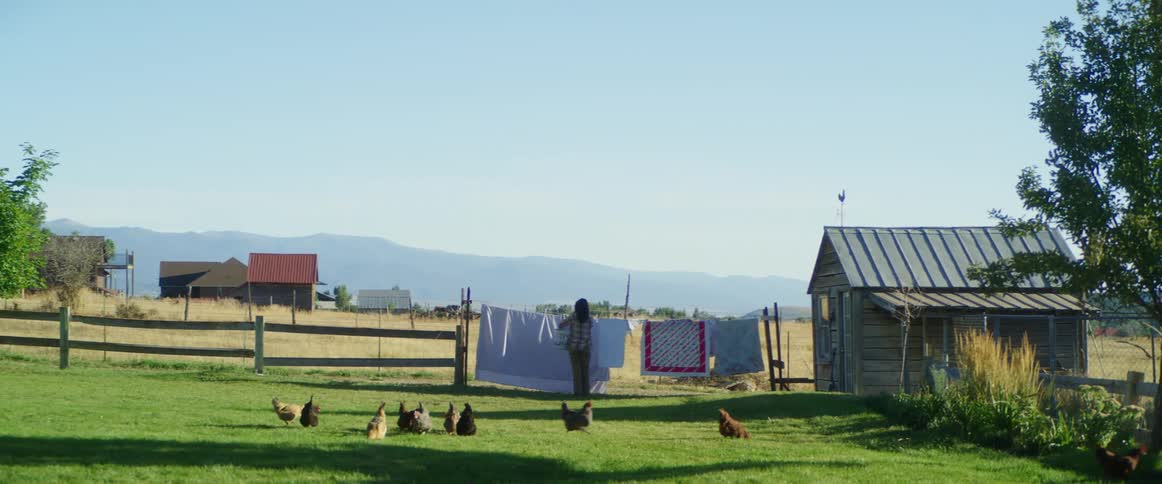A woman hangs clothing on a clothes line outside with chickens running round the rural fenced in yard