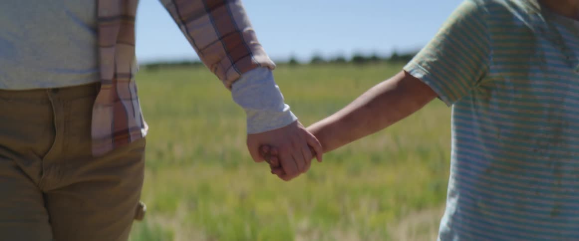 A brother and sister walk across a grass field holding hands and then the little boy lets go and runs