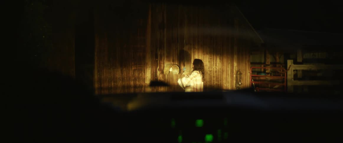 A vehicle pulls up and stops in front of a barn where a woman stands holding something in her arms