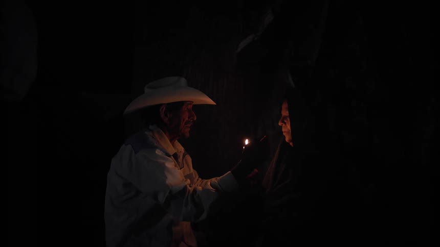 A man in a cowboy hat smiles as he holds a lighter up to a woman in a head scarf