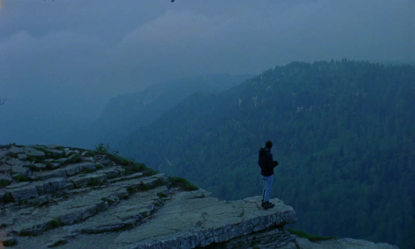 A photographer walks to the edge of a mountain cliff and raises his camera to take a photograph of the foggy landscape below