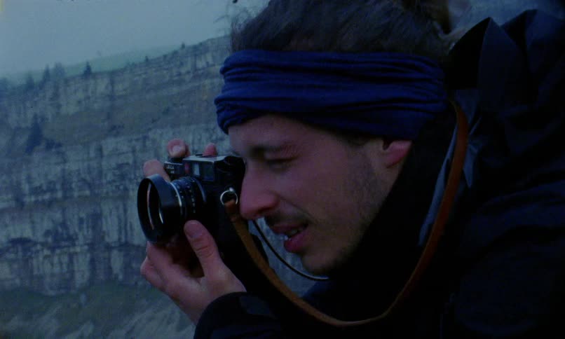 Photographer stands outdoors on windy day taking pictures with mountain cliff as backdrop
