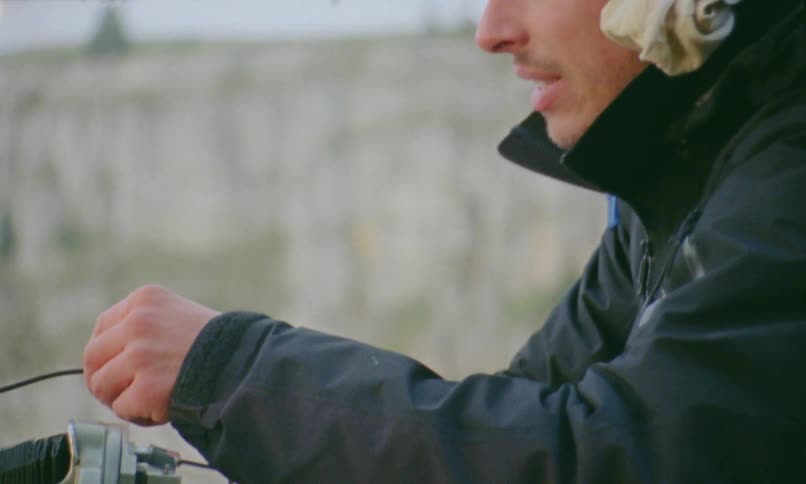 Young man wearing a thick jacket sets up a vintage camera on a tripod outdoors in a windy day