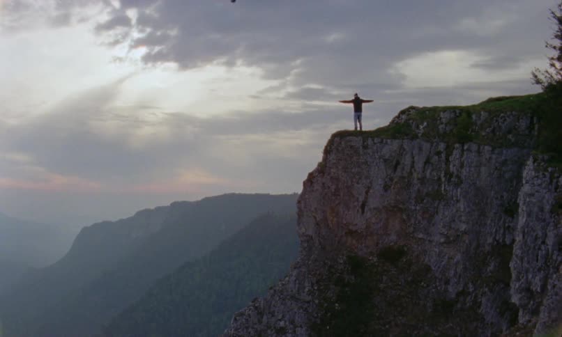 Man with a coat jeans and a pair of boots stands at the edge of a cliff on a cloudy day