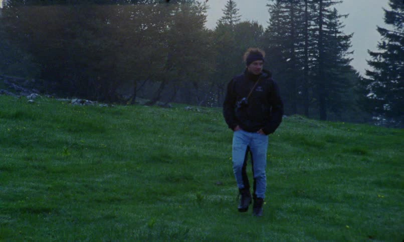Man with blue pants a pair of boots a black coat and a black headband walks down a grass hill