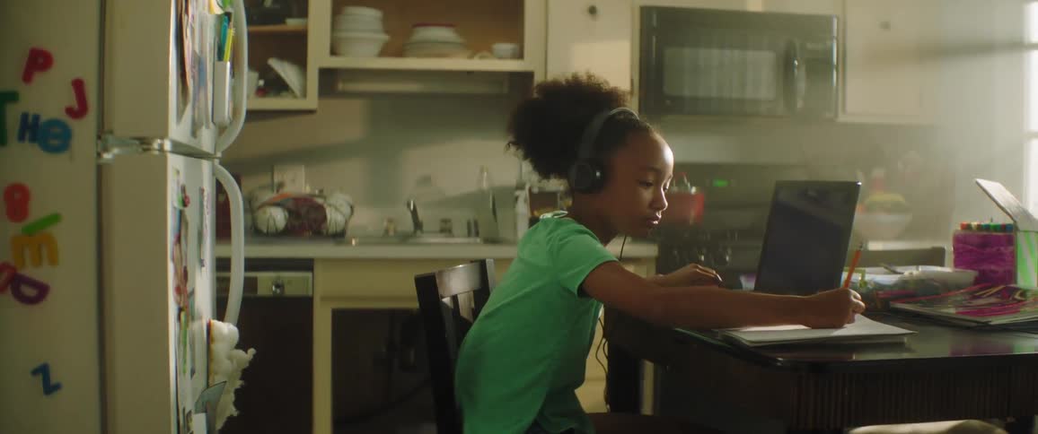 Young girl sitting at the table in the kitchen of her home places a set of headphones over her ears and begins to write with a pencil onto the paper sitting next to a laptop