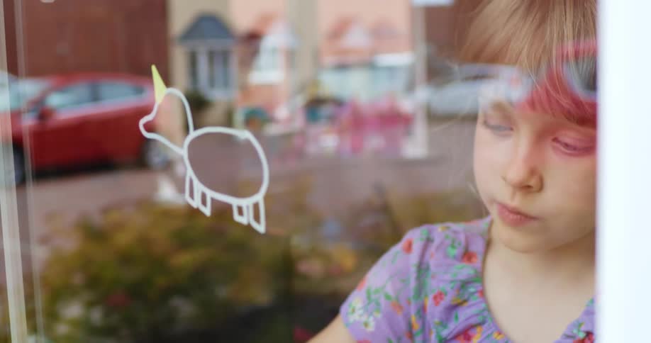 A young girl uses a yellow marker to add a horn to a unicorn drawing on her window