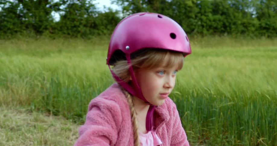 Blonde girl with a pink helmet and a jacket looks around and starts cycling on a dirt road in the countryside