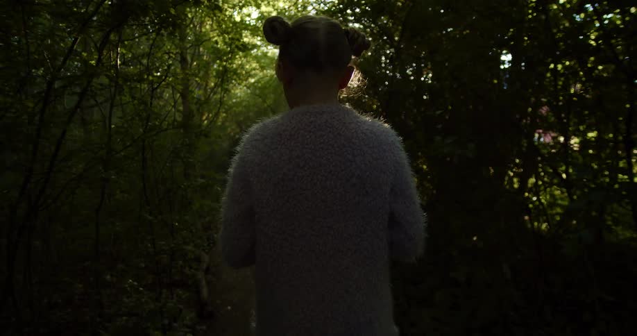 Blonde girl with a grey pullover walks along a track as the sun shines through trees in the forest
