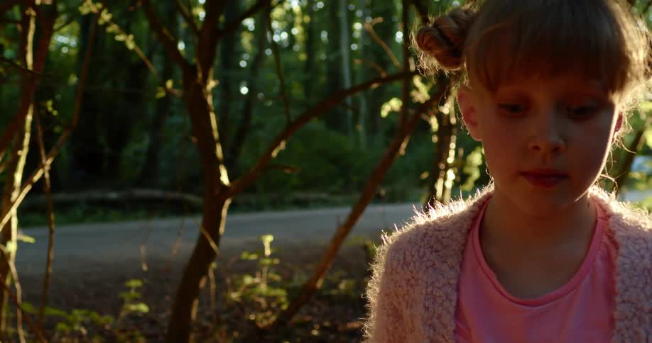 A girl walks through a woodland next to a road as sunlight shines through the foliage onto her back