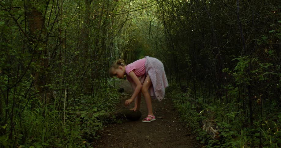A young girl walks a dirt path in a heavily wooded area and bends down to tear up the soil