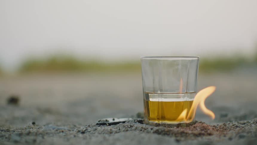 Flames rise from the bottom of a beverage glass sitting on the sand