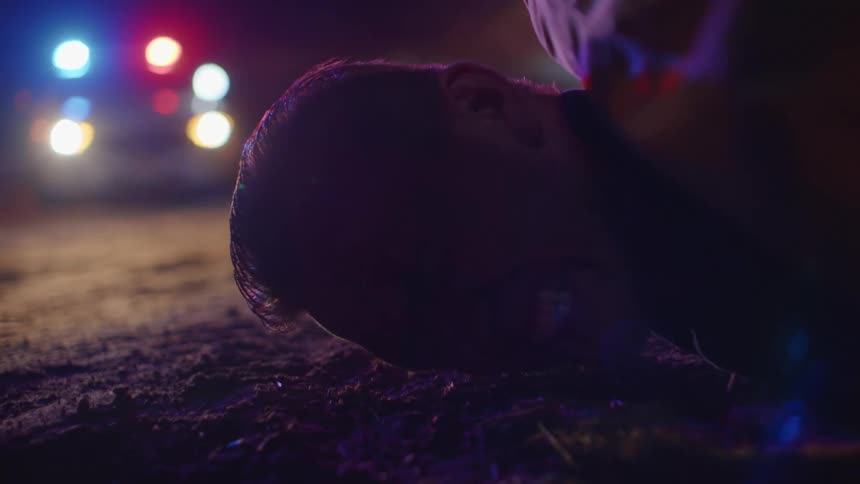 A man lies on the ground crying as police lights flash in the background