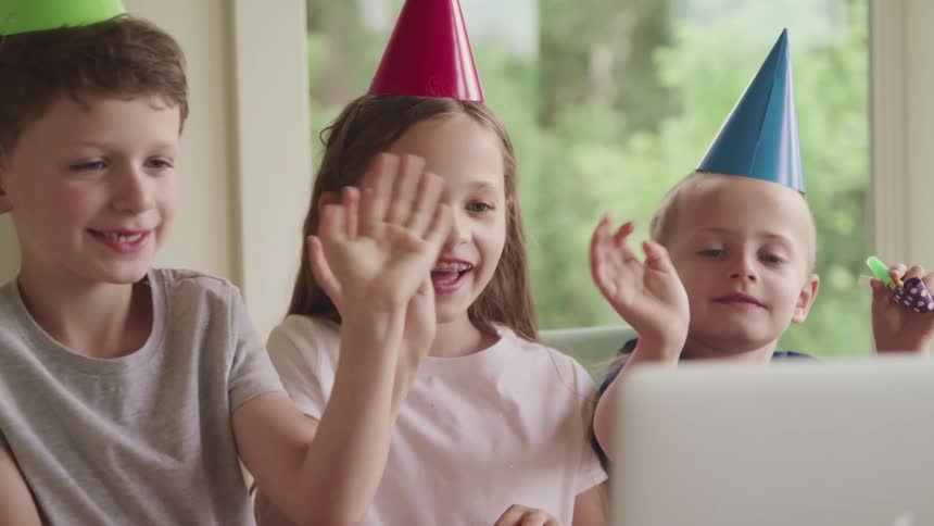 Two boys and a girl wearing colorful party hats wave smiling at a laptop screen at home