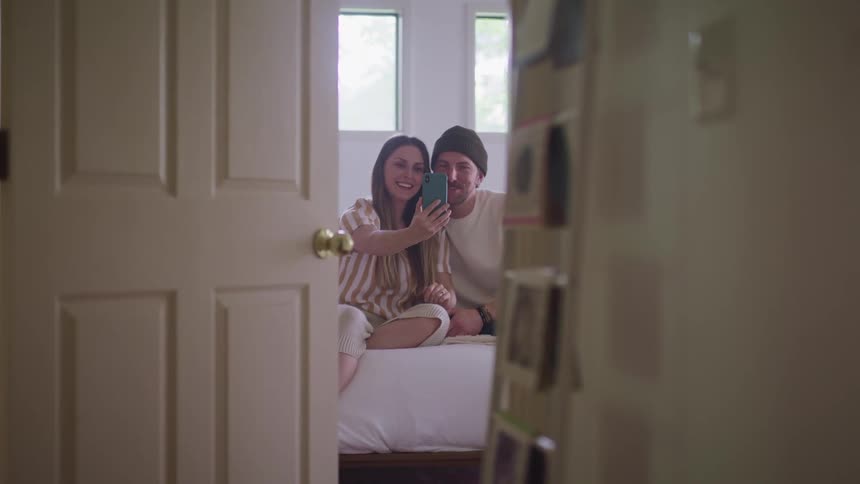 Woman with a striped shirt and brown hair and man wearing a dark green beanie and a white sweater sitting on the bed make a video call and wave to somebody on a smart phone in the bedroom
