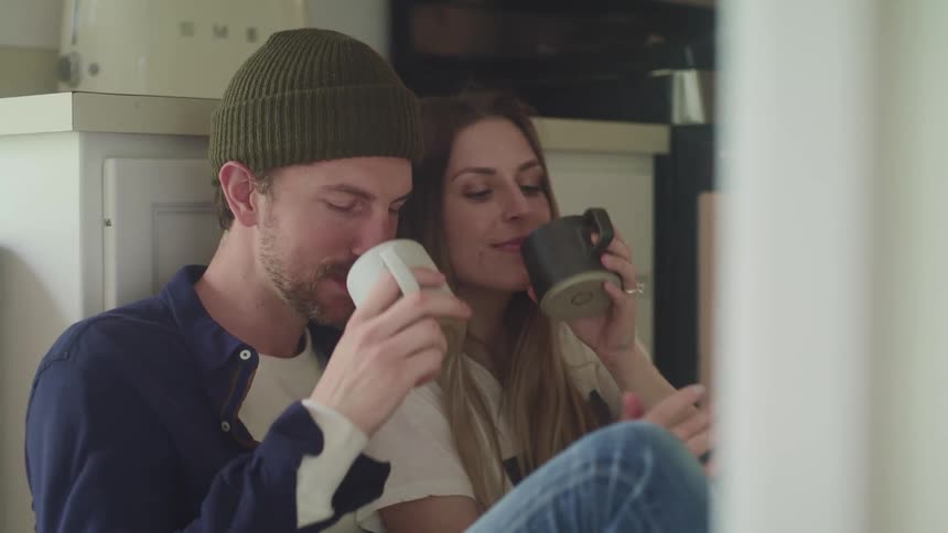 A man and woman sit against a cabinet on the floor and lean against one another as they drink coffee and laugh