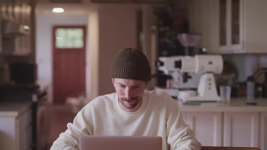 A man is sitting in a kitchen and using a laptop