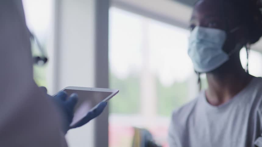 Female patient wearing a face mask sits in front of doctor as they type medical information onto a tablet