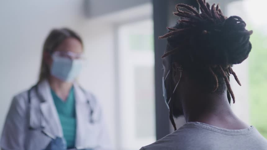 A male patient sits in an office as a female doctor stands across from him writing on a clipboard