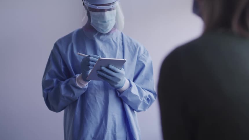 A person wearing a disposable medical gown and lots of protective equipment interviews a woman