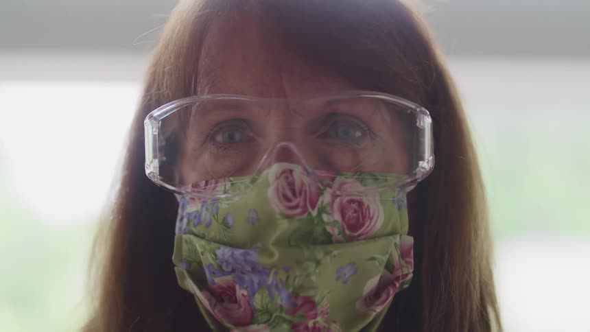 A woman in a medical mask and safety goggles looks into the camera